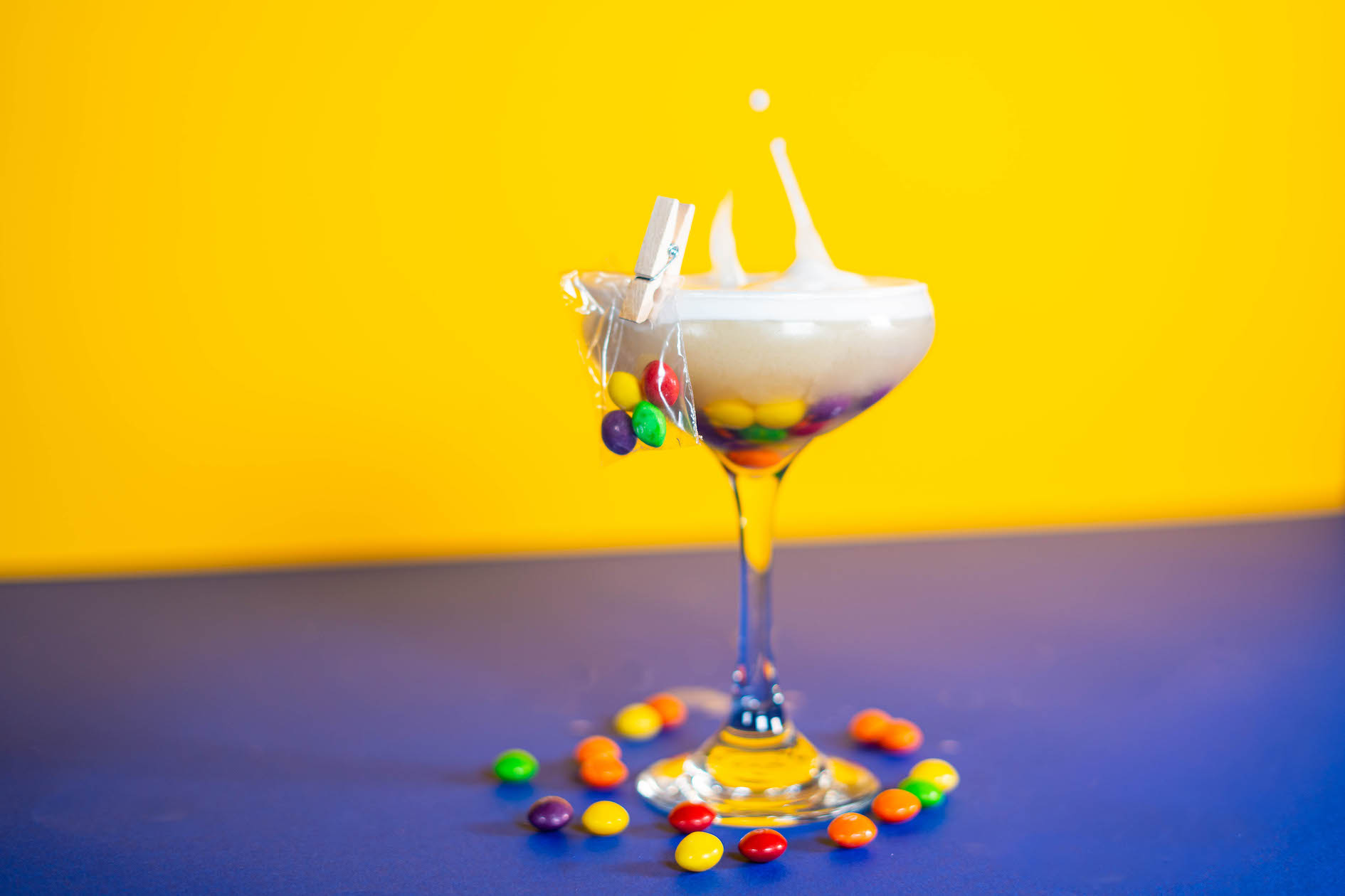 a cocktail with skittles in a packet pinned to the glass and skittles all around it