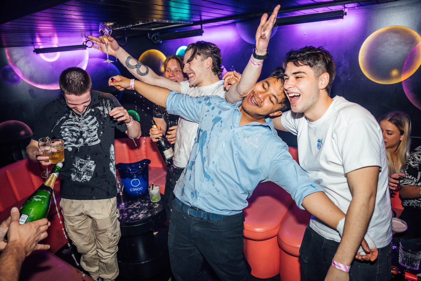 a group of men dancing in a VIP booth, having a bottle of champagne sprayed over them