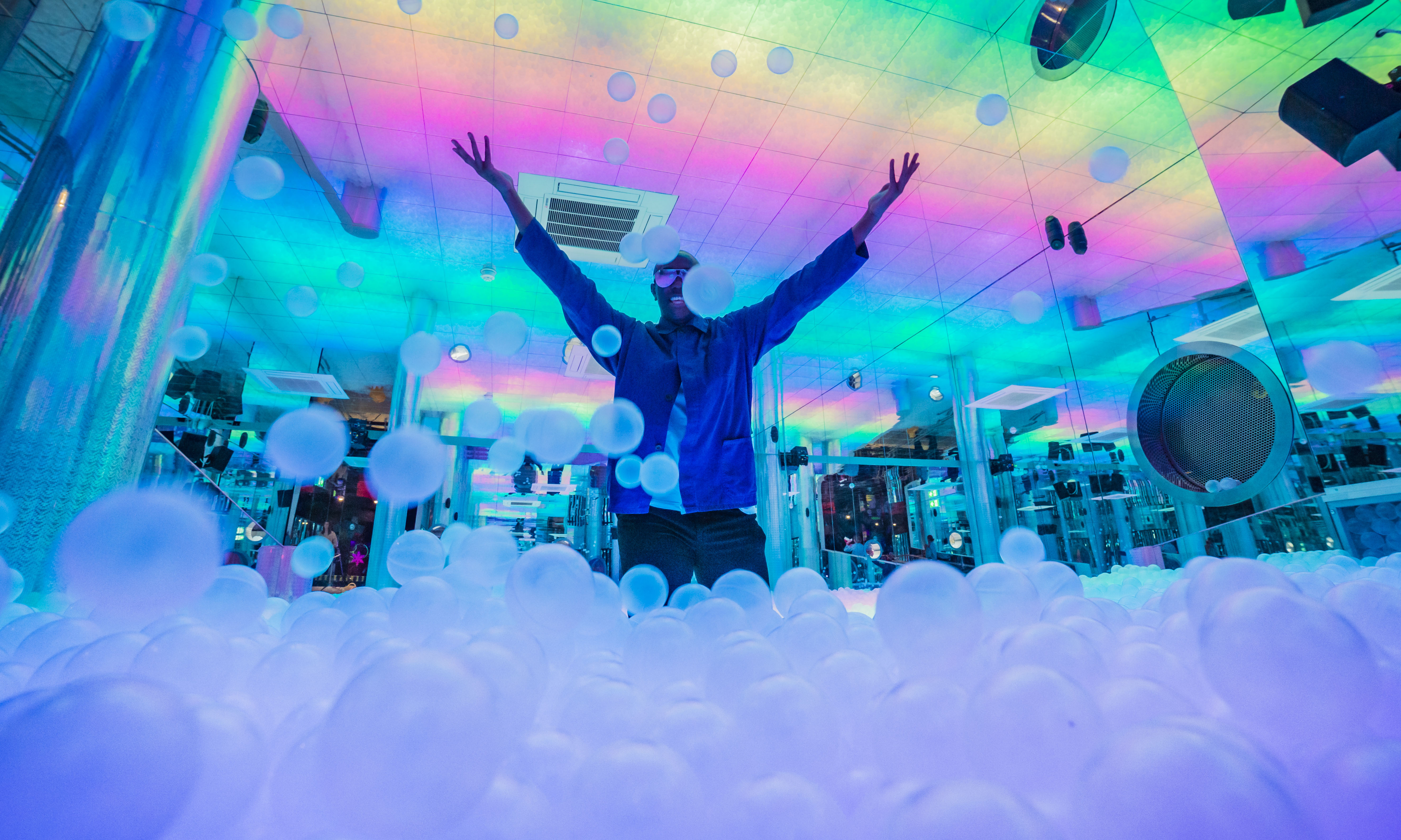 Man in blue shirt in rainbow ball pit throwing balls up
