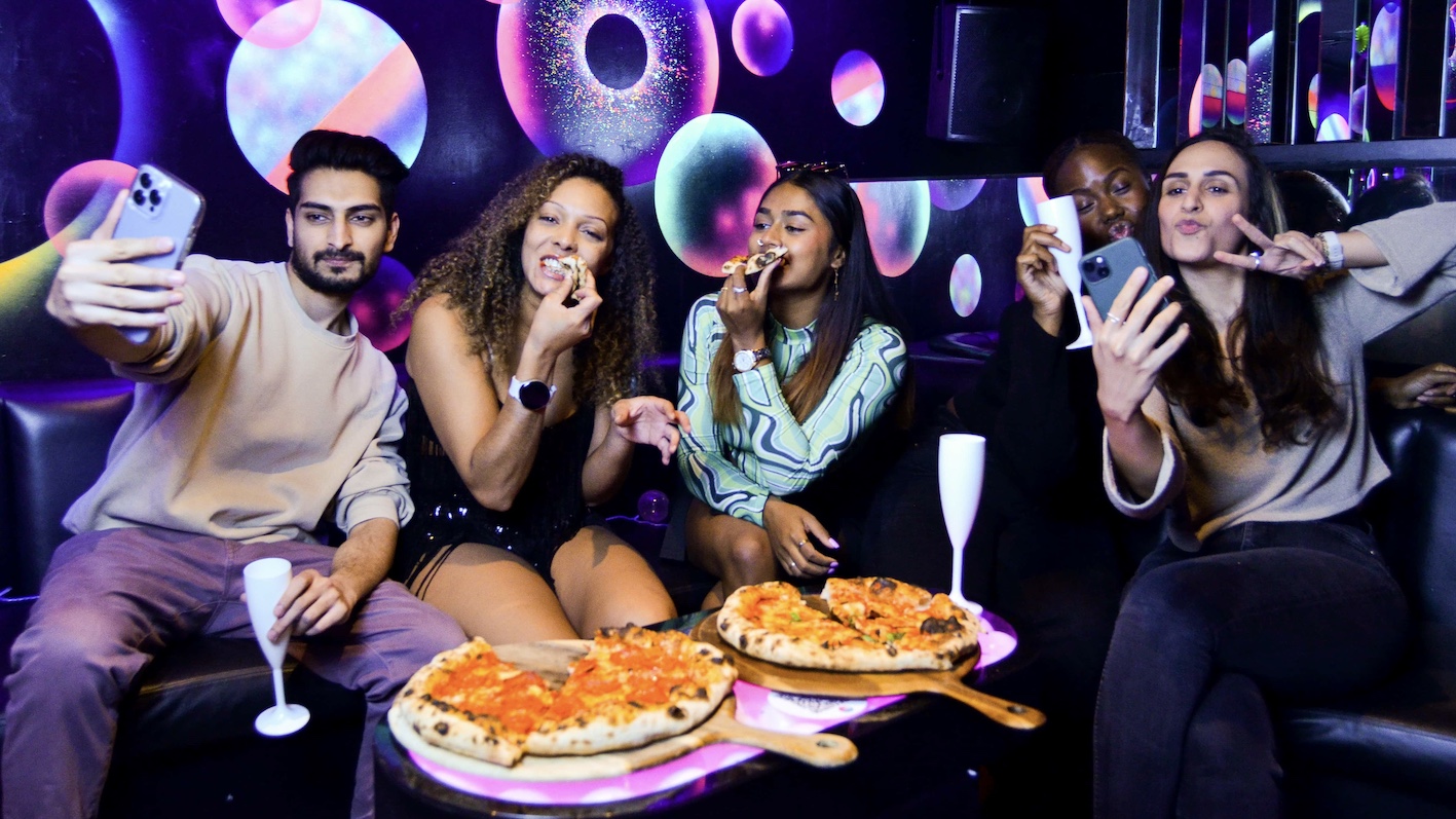 a group enjoying pizza and cocktails in a VIP booth, taking selfies