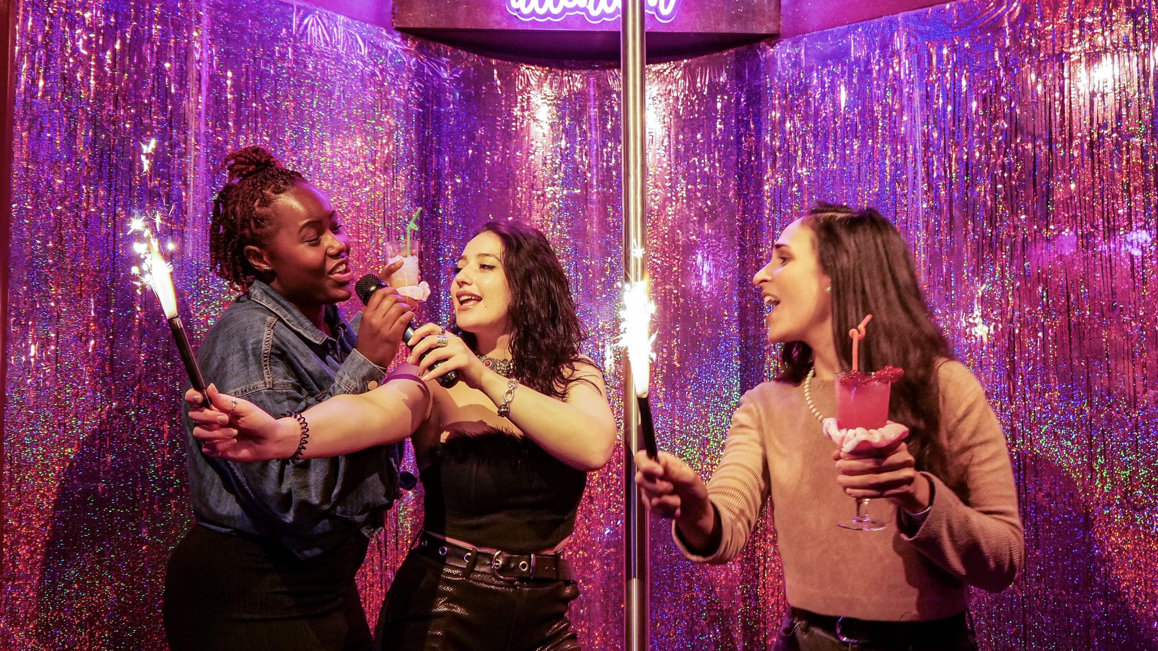 three women singing in a. karaoke booth, holding sparklers and cocktails
