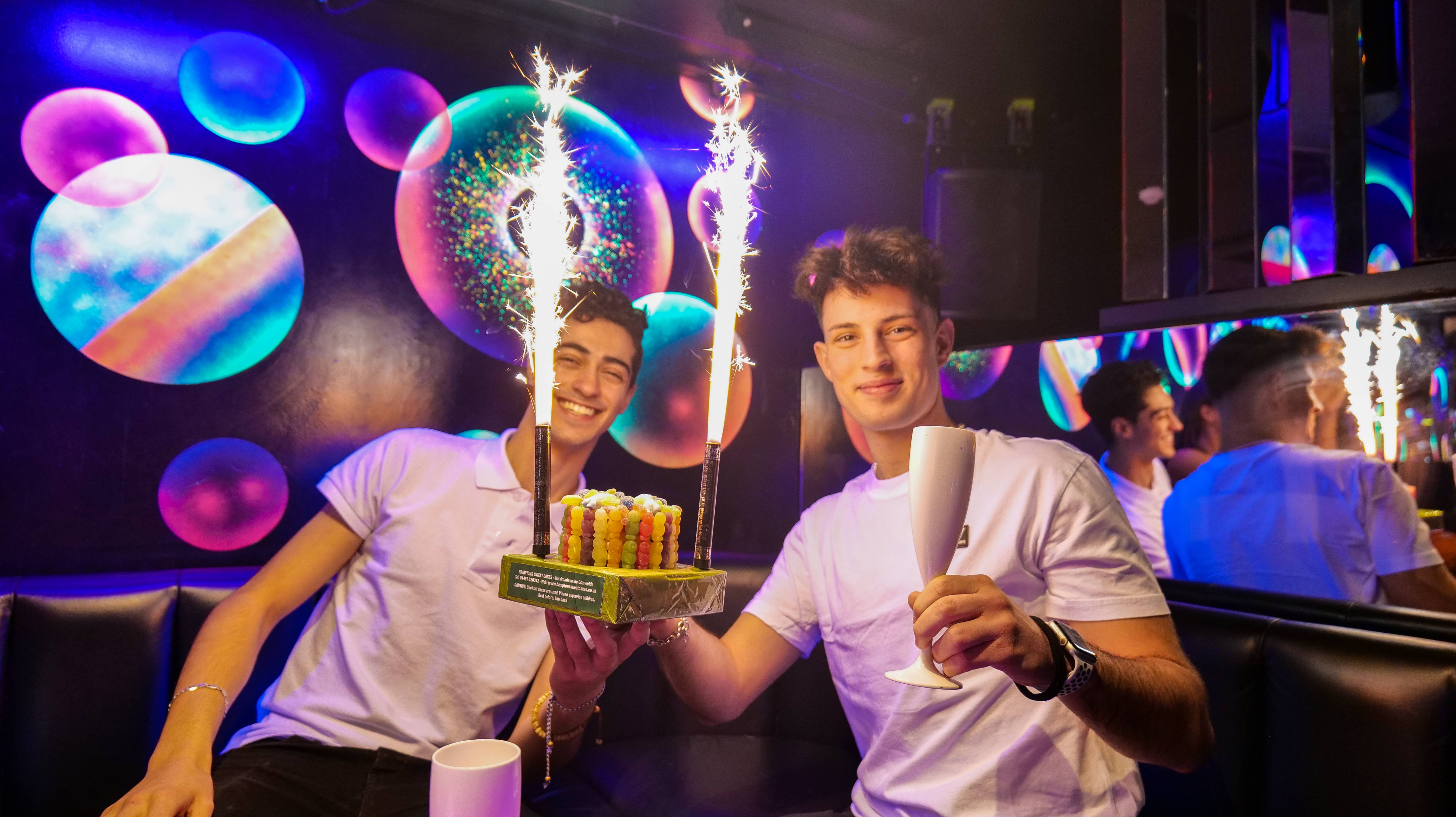 two men holding a birthday cake with sparklers drinking prosecco