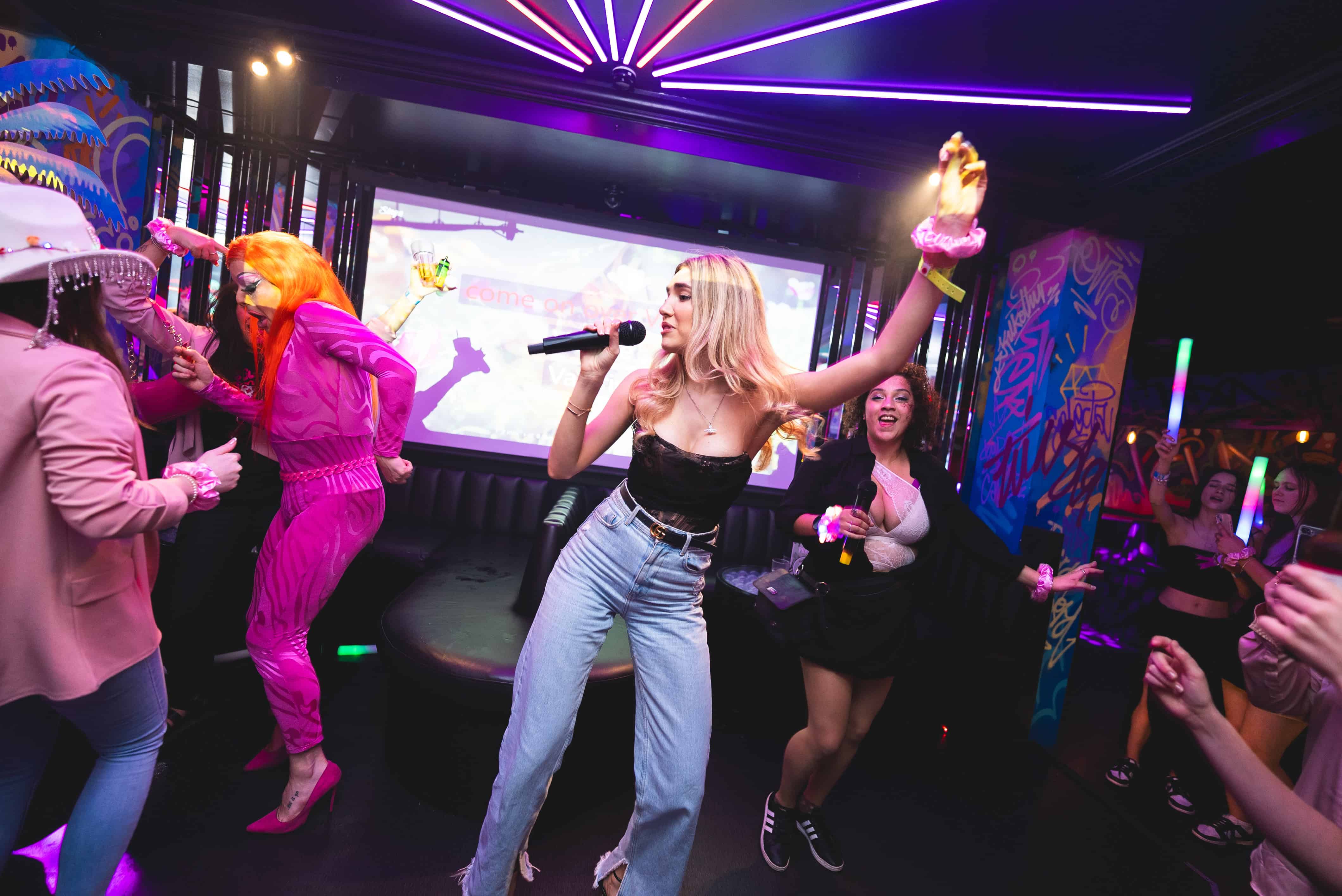 three women singing in a. karaoke booth, holding sparklers and cocktails