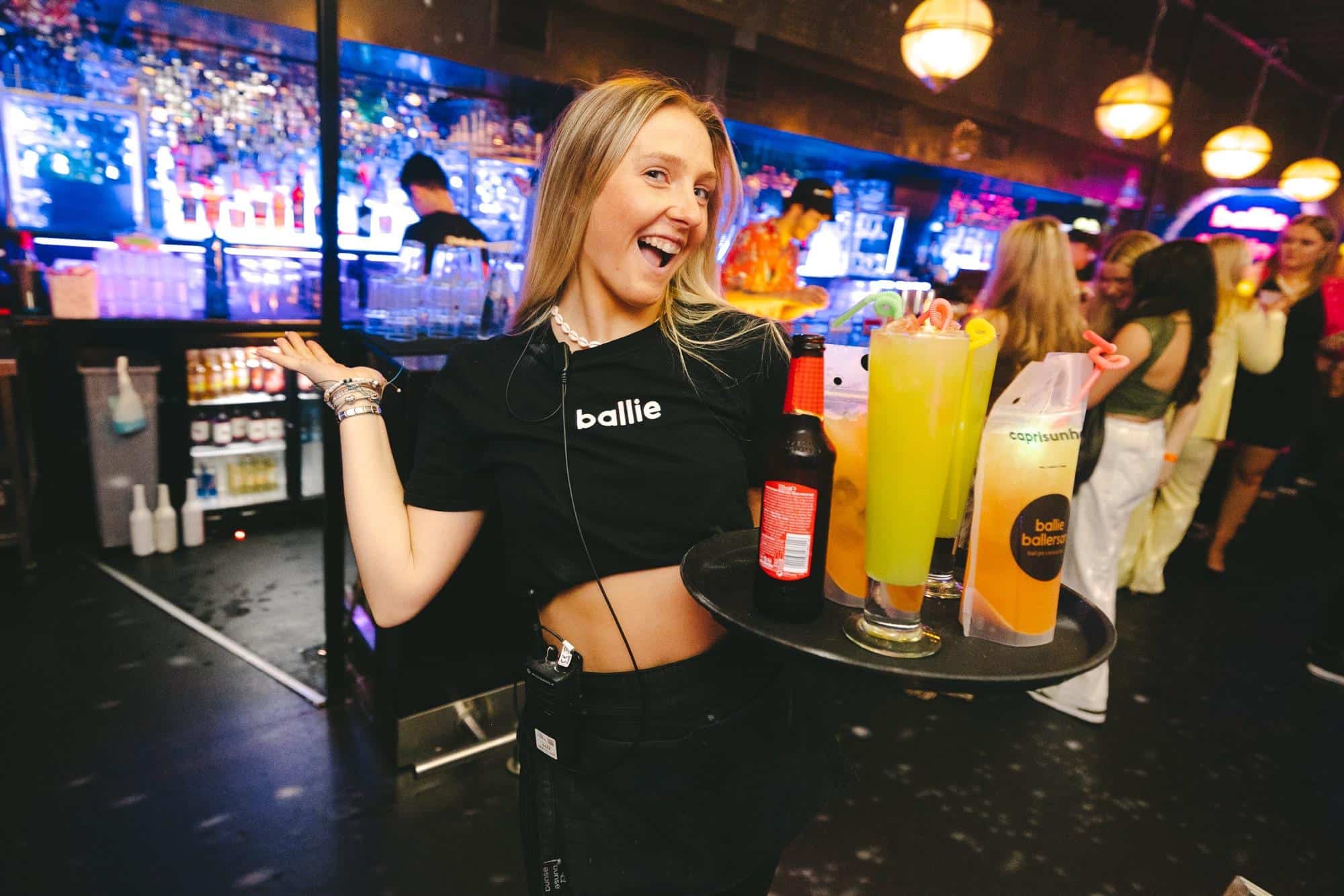 Vibrant nightclub scene at Ballie Ballerson with a smiling waitress holding drinks and a lively crowd enjoying the neon-lit atmosphere.